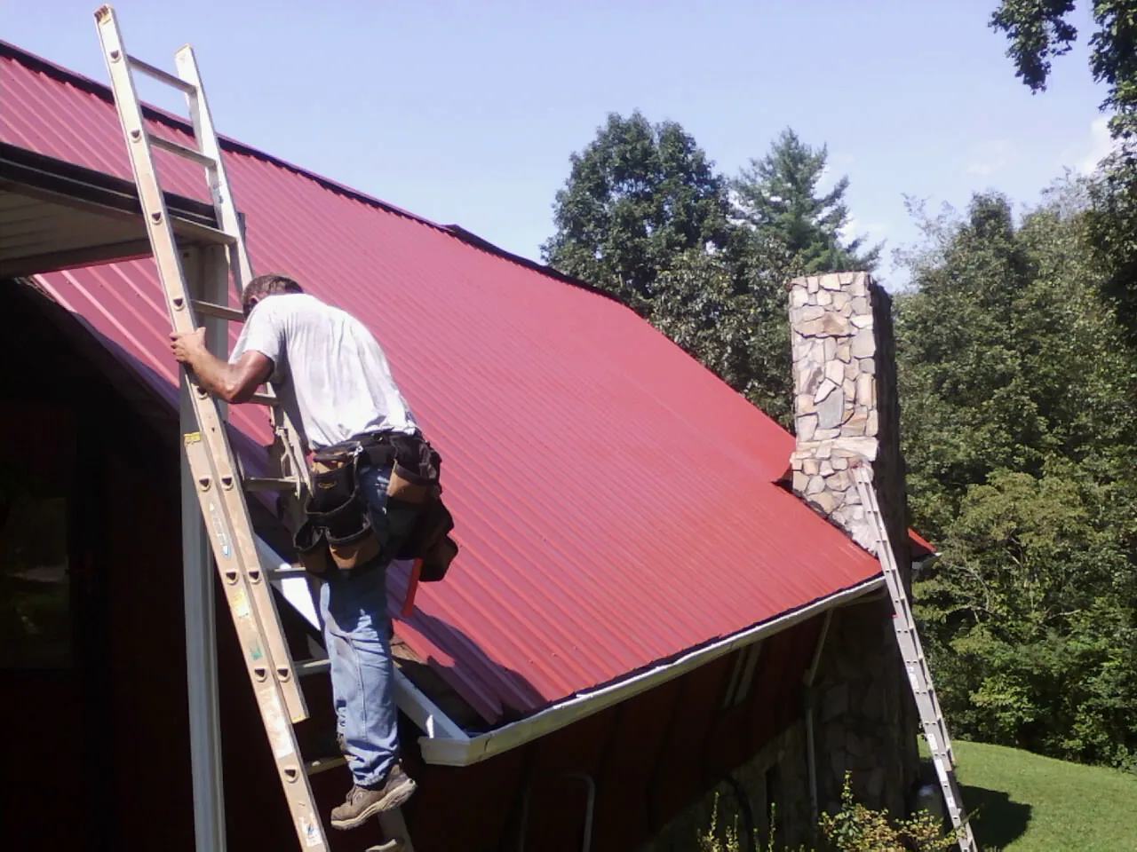 Roofer on a ladder beside a red metal roof and stone chimney — Soffit & Fascia Repair by Roofing Brothers in Sharon Springs